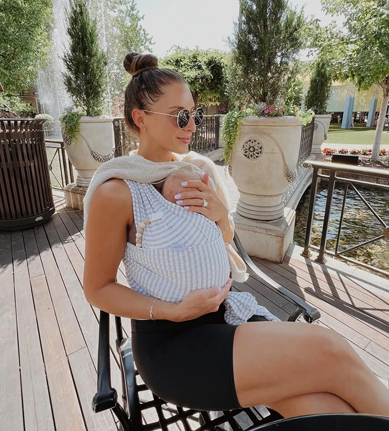A woman in sunglasses sits on a patio holding a baby in a striped sling carrier.