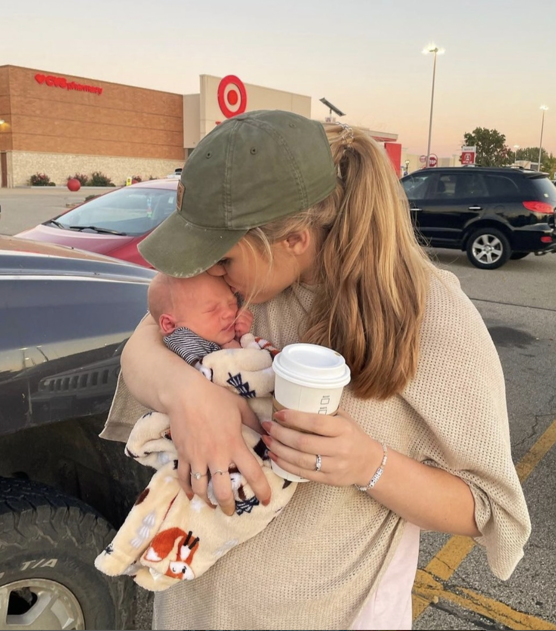 Person holding a baby wrapped in a blanket, standing in a parking lot.