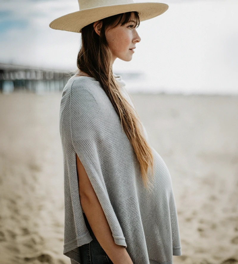 A pregnant woman in a straw hat and grey knit Cocoon poncho stands on a beach looking to the side.