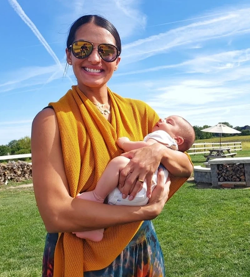 A smiling woman in sunglasses holds a baby outdoors in a grassy field on a sunny day.