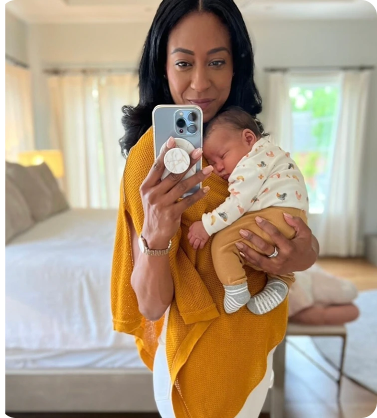 Woman taking a mirror selfie while holding a sleeping baby in a cozy room.