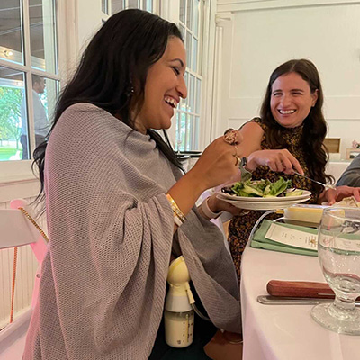 Two women smiling and eating at a dining table indoors.