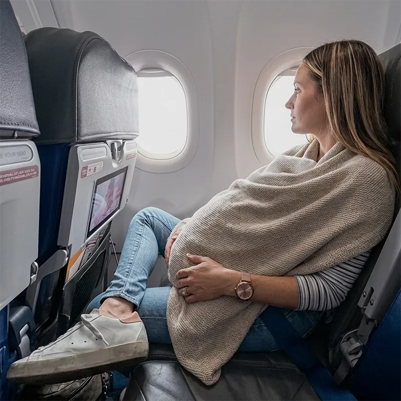 mom with straight blonde hair looking out the window of an airplane with a sleeping baby under a cocoon nursing cover