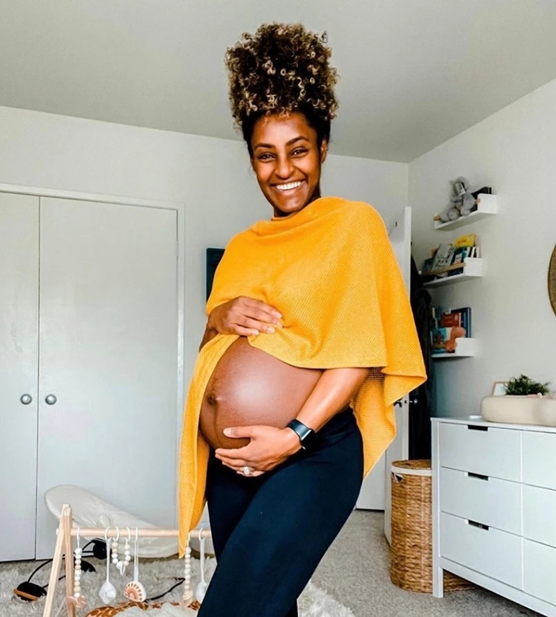 A smiling pregnant woman in a yellow shawl holds her bare belly while standing in a room.