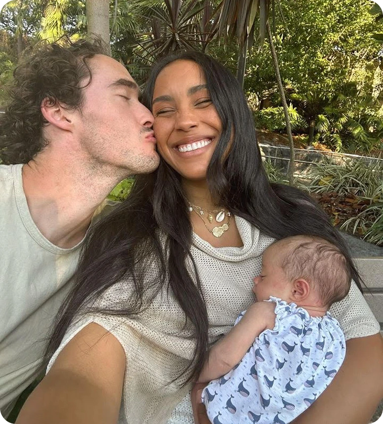 A couple smiling with a baby outdoors in a garden setting.
