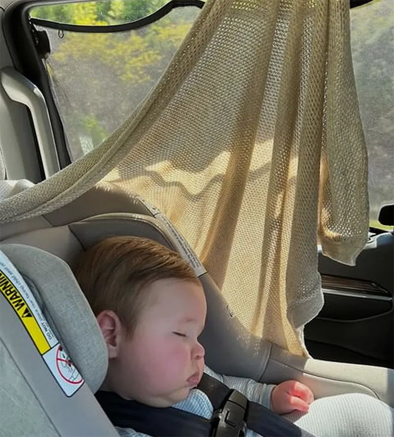 A baby sleeps peacefully in a car seat, partially shaded by a knit blanket hung on the window.
