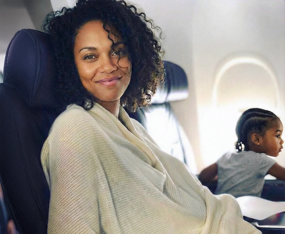 A woman with curly hair smiles while sitting on an airplane next to a young child.