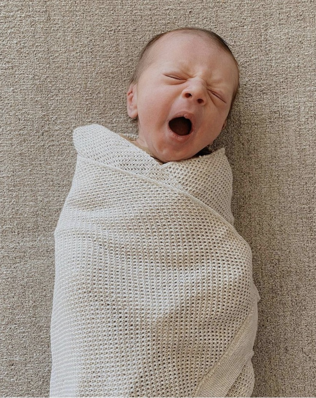 Yawning baby wrapped in a beige blanket on a textured surface.