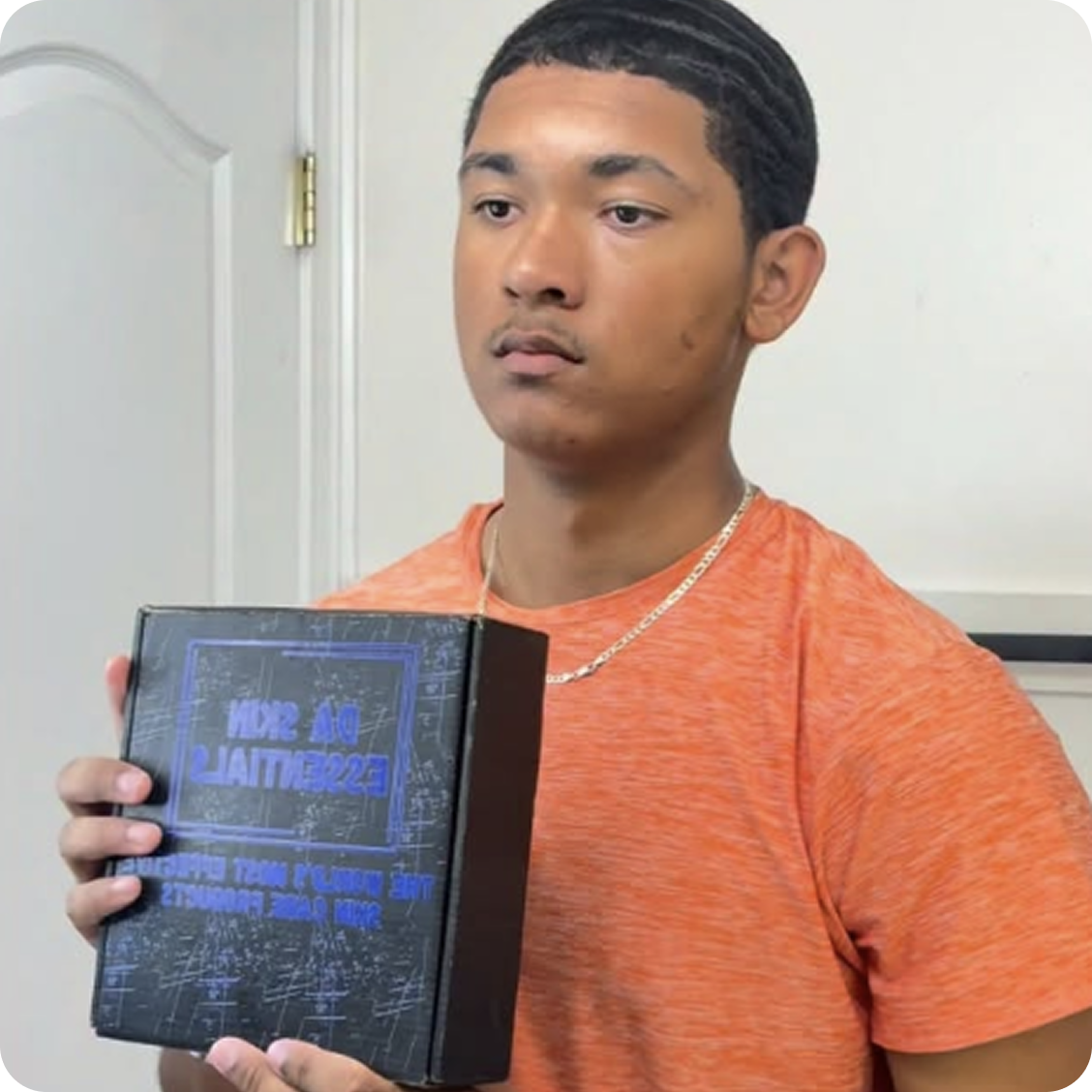 A young man in an orange t-shirt holds a black box with blue text.