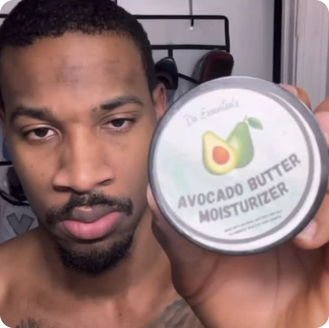 A shirtless man with a goatee holds up a round tin of Avocado Butter Moisturizer to the camera.