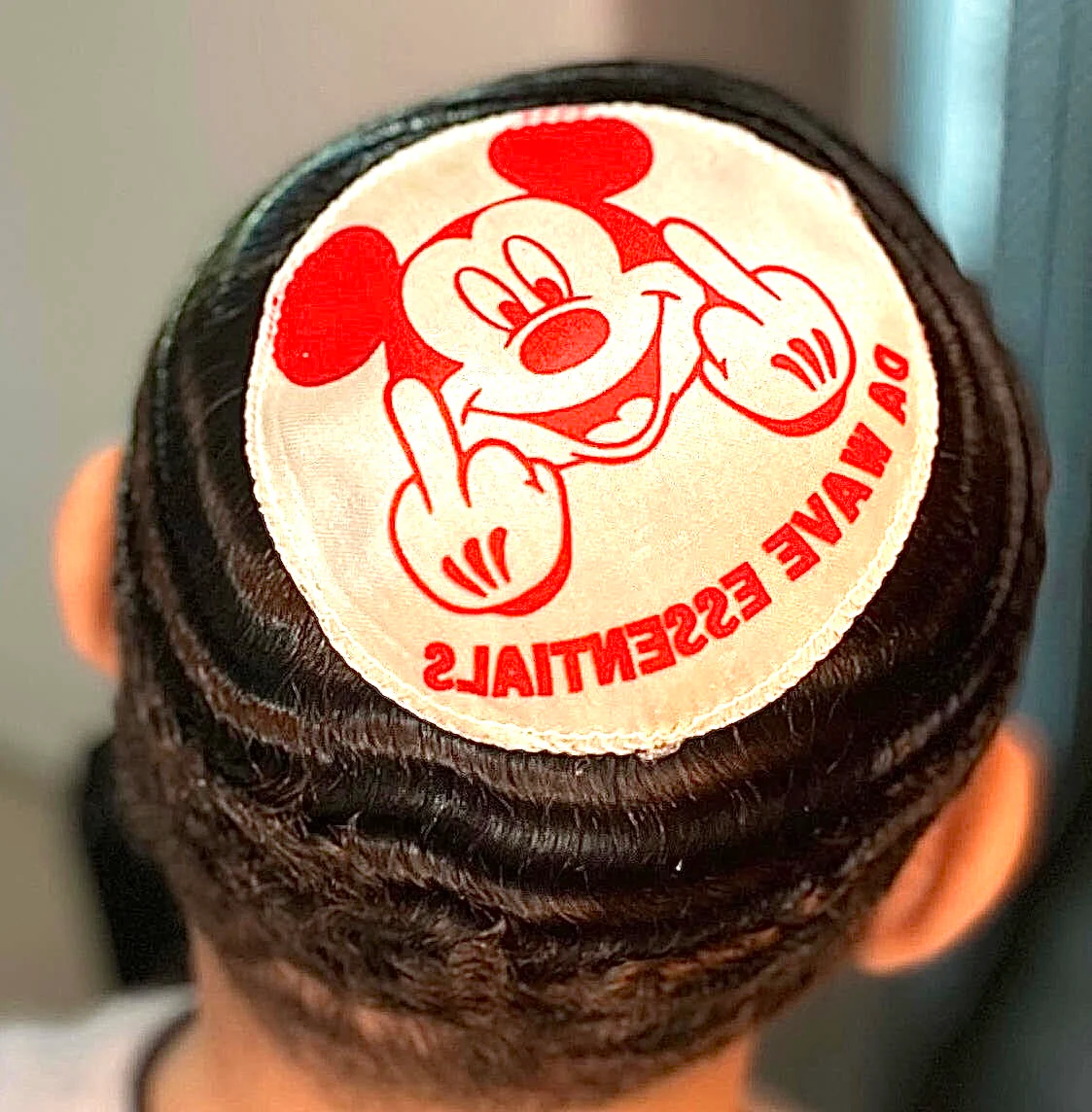 The back of a head with waved hair wearing a patch of a cartoon mouse giving the middle finger.