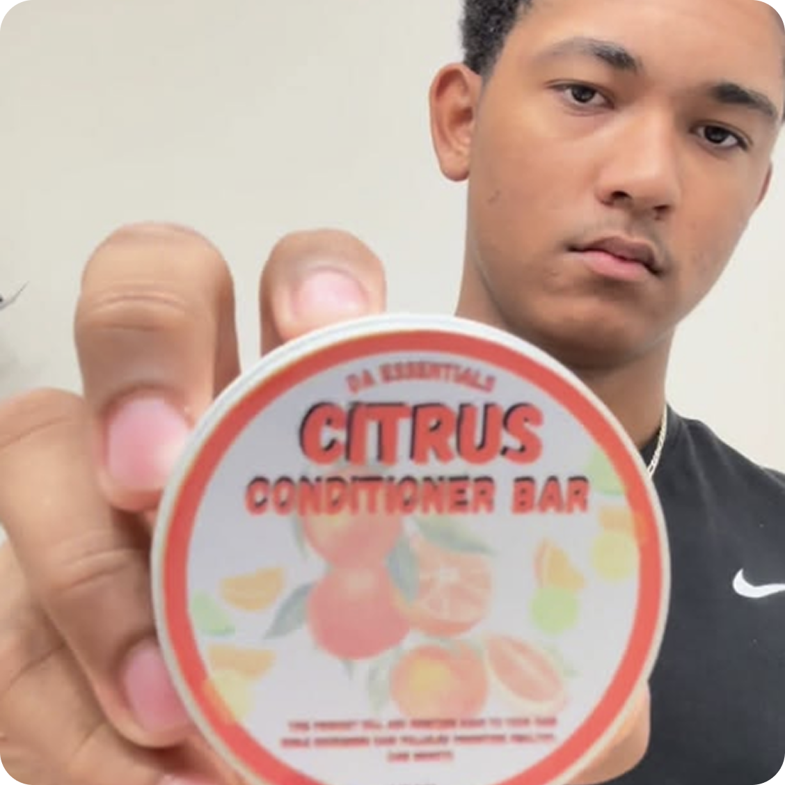 A young man holds up a round tin of DA Essentials Citrus Conditioner Bar to the camera.