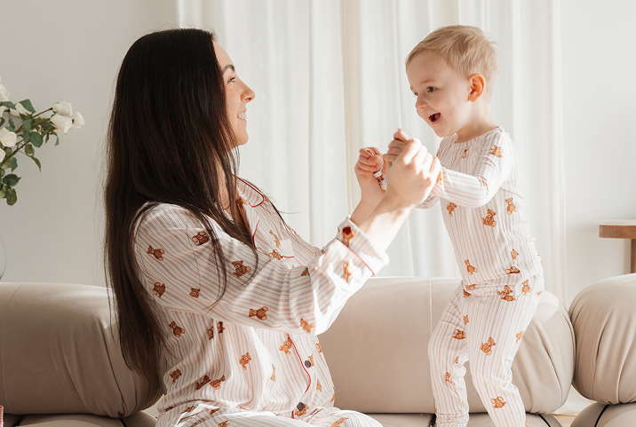 A mother and child in matching teddy bear pajamas smile and play on a couch.