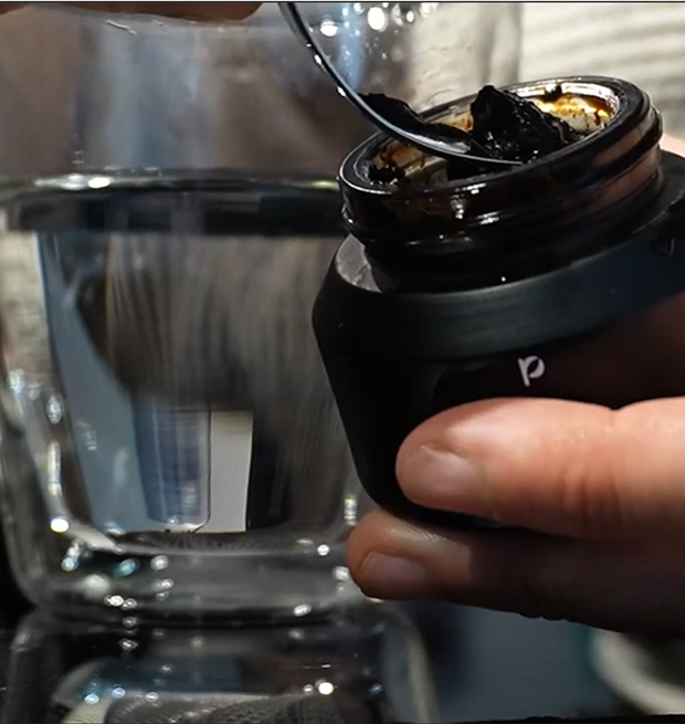 Hand holding a jar with dark substance near a glass of water.