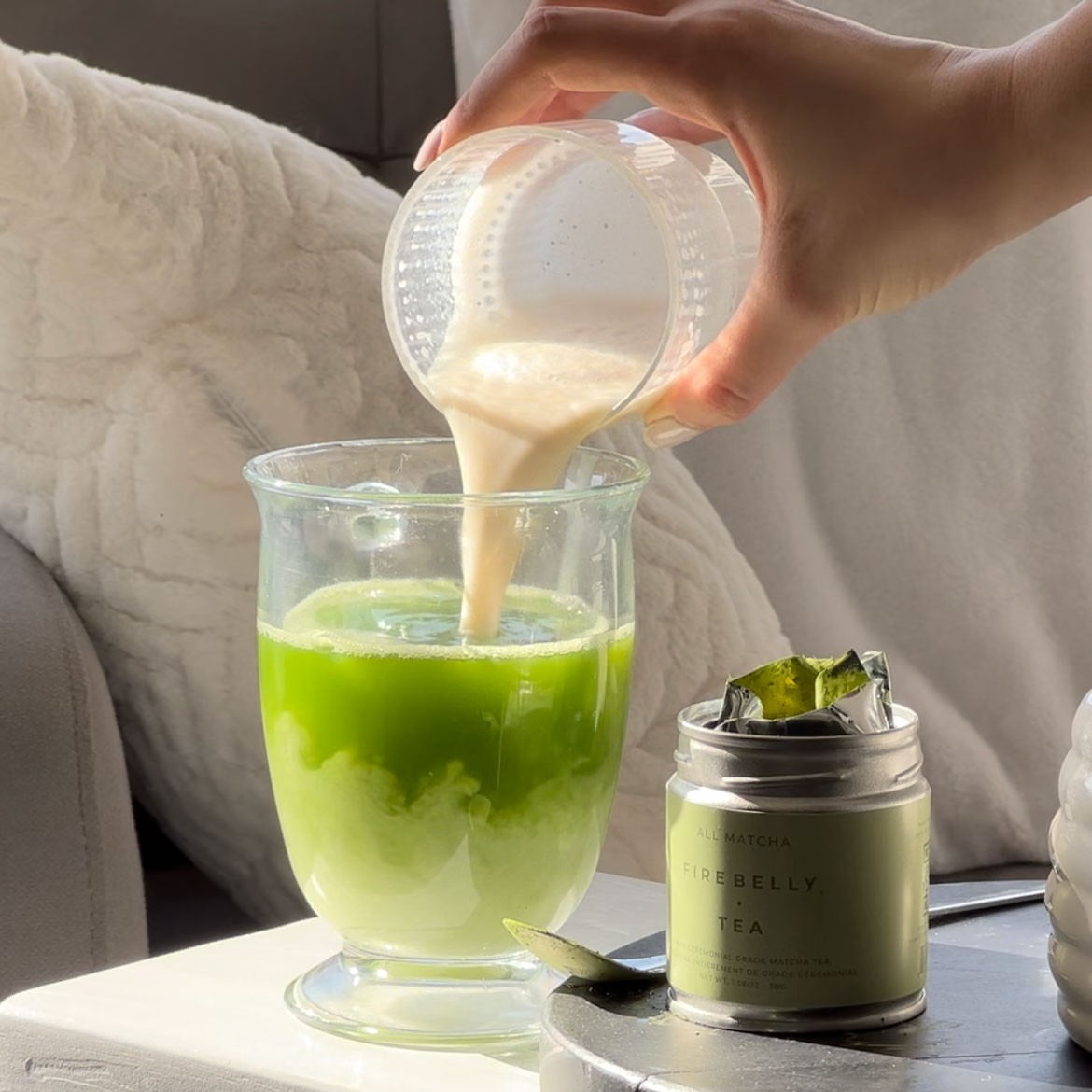 Hand pouring milk into green matcha tea beside a canister on a white surface.