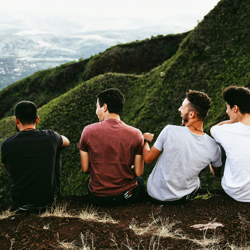 Four friends sitting together, laughing on a grassy hillside with a stunning view.