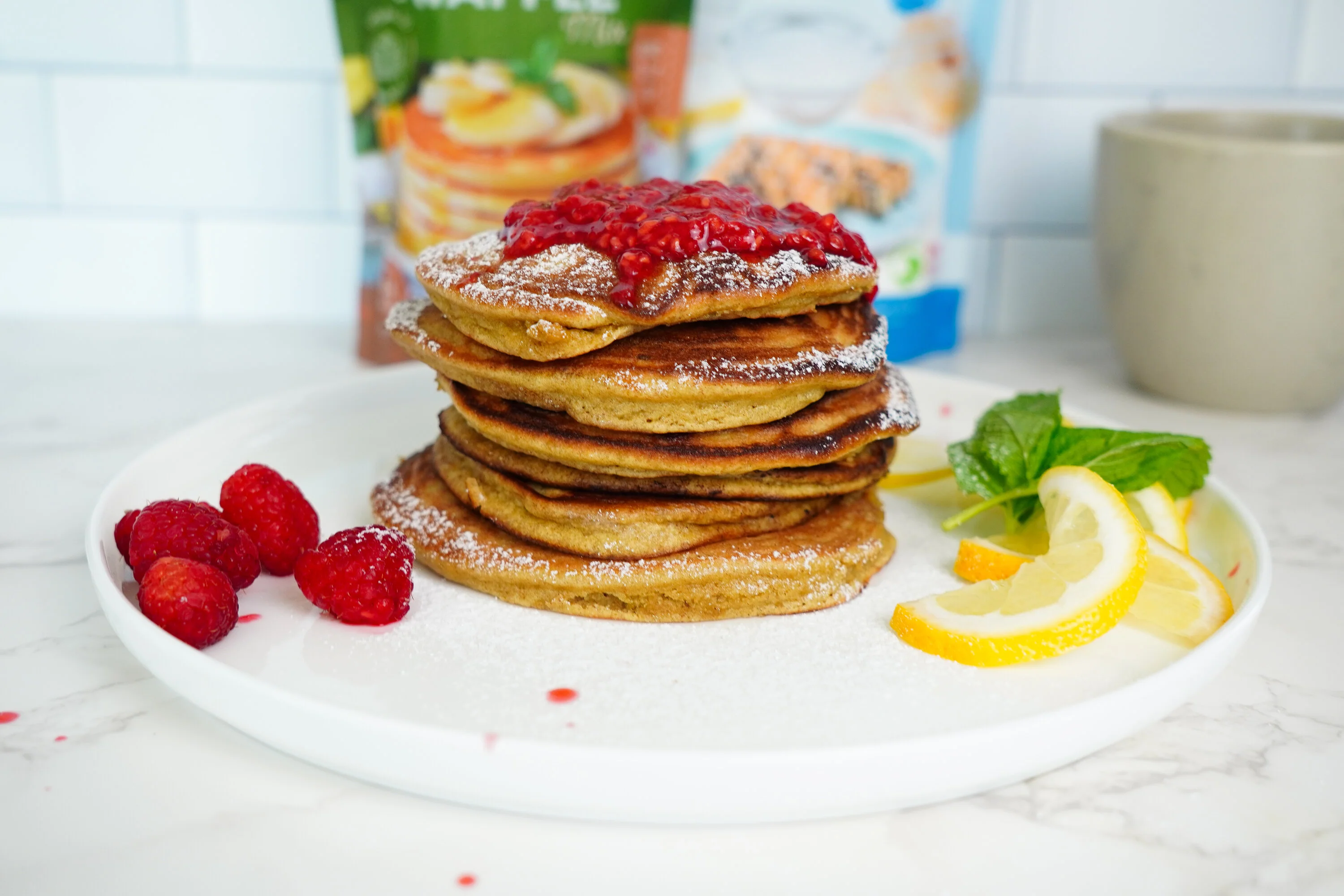 Stack of pancakes with raspberries, lemon slices, and mint on a white plate.