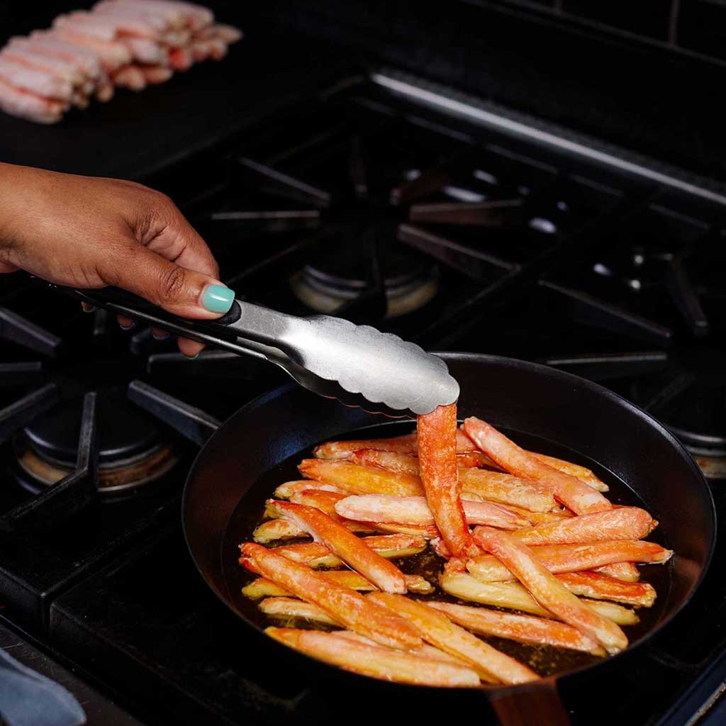 A hand uses tongs to cook imitation crab meat in a black pan on a stovetop.