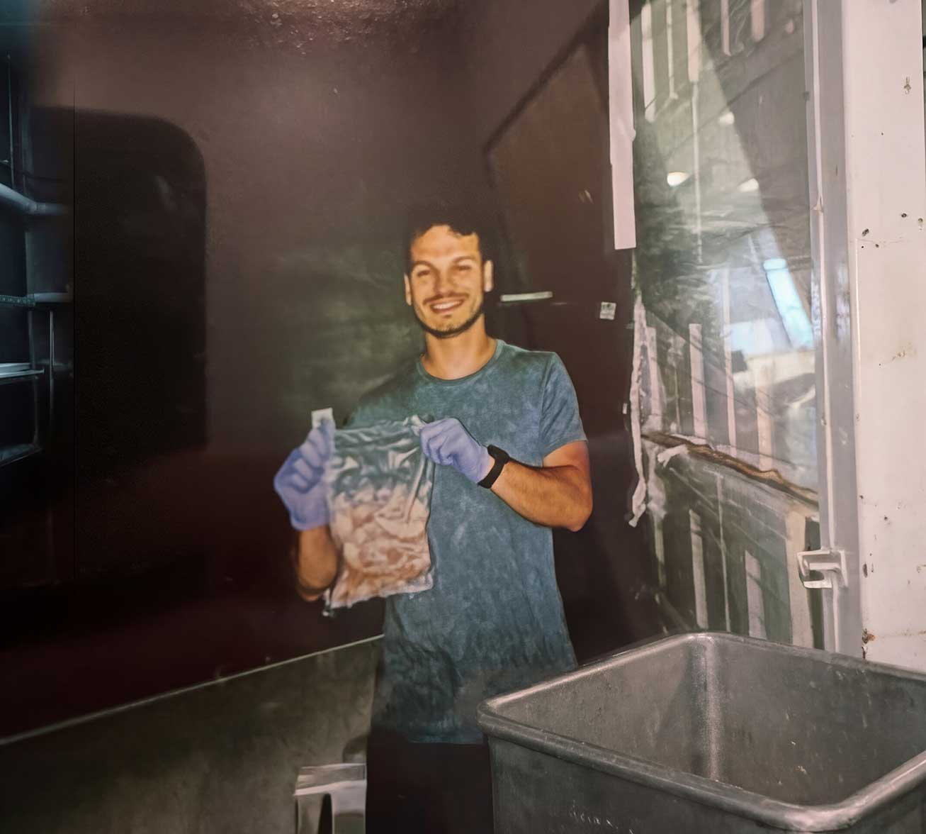 A smiling man wearing blue gloves holds up a clear plastic bag filled with food.