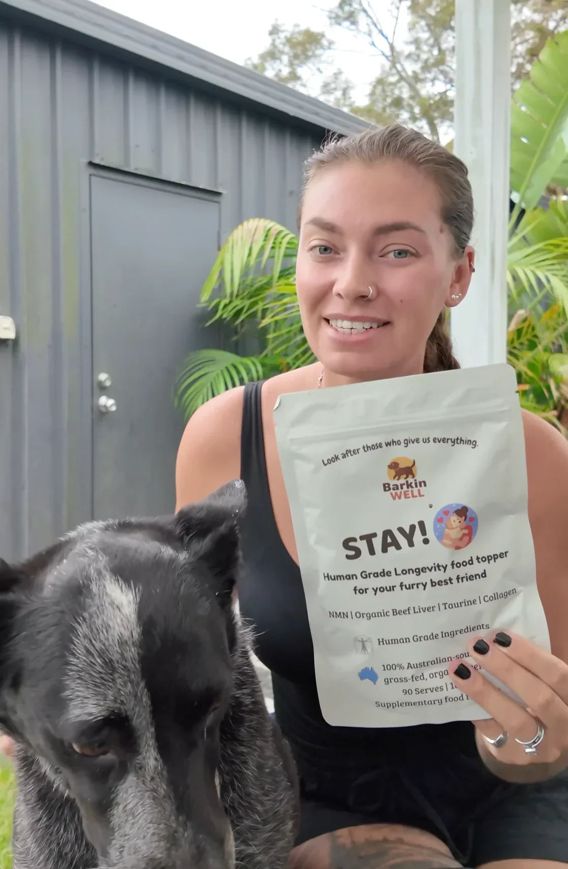 A smiling woman holds a pouch of Barkin Well dog food topper next to her black and white dog.