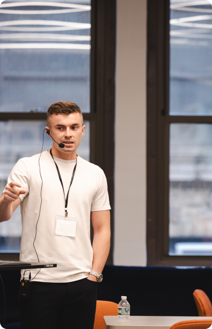 A man in a white t-shirt with a headset microphone speaks while gesturing with his right hand.