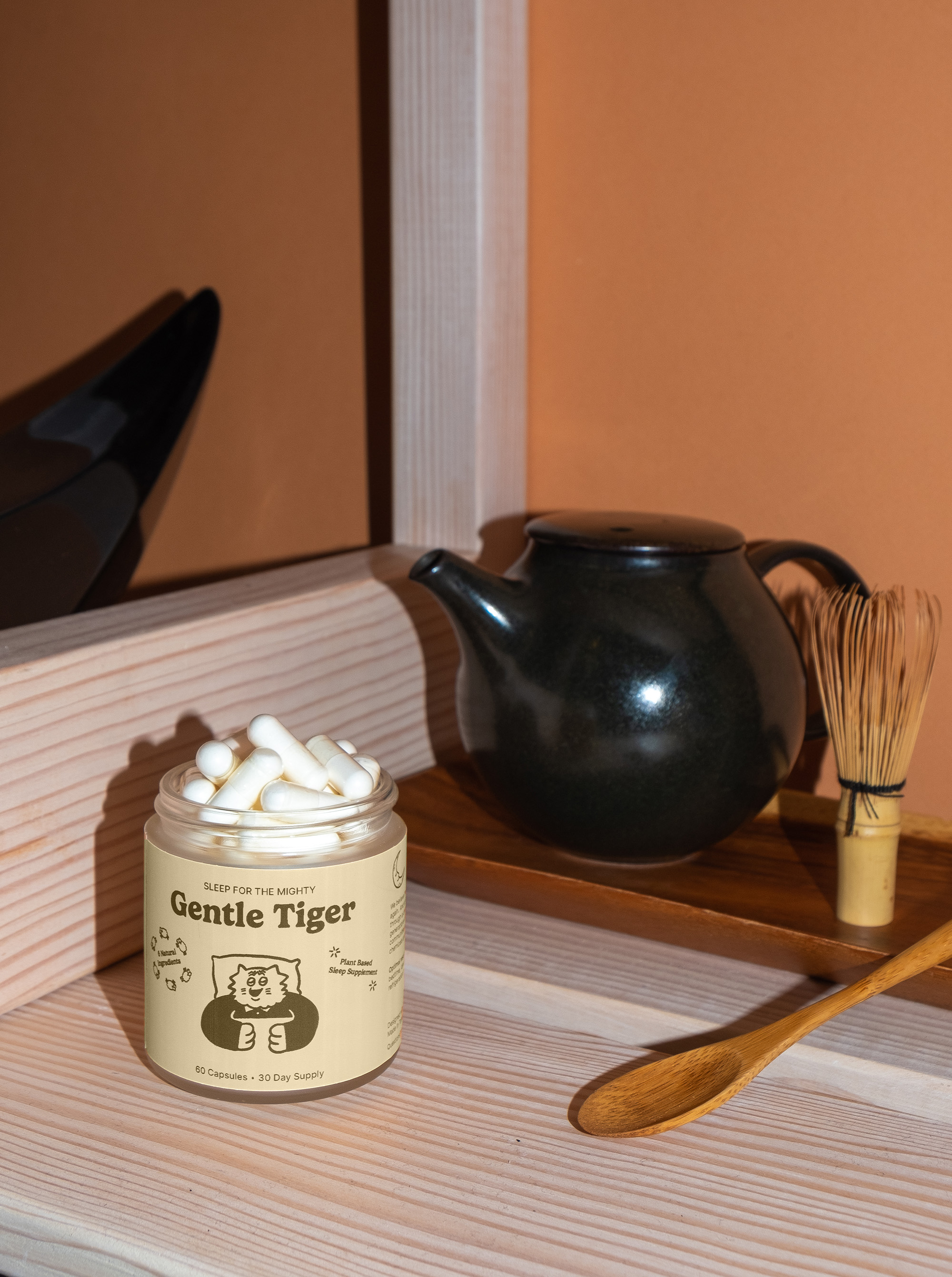 Jar of capsules labeled 'Gentle Tiger' next to a teapot and whisk on a wooden surface.