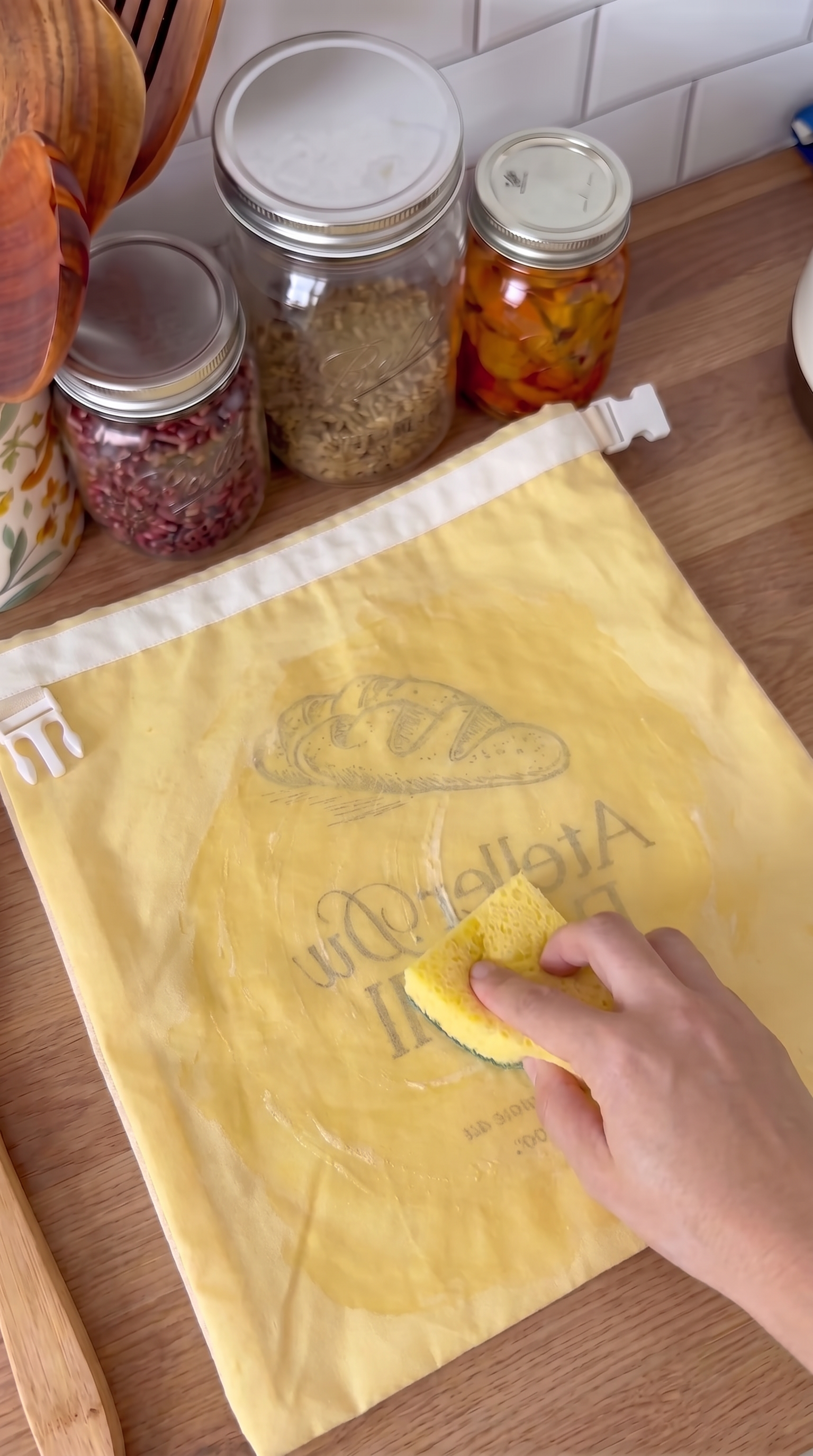 A person's hand wipes a yellow reusable bread bag with a sponge on a kitchen counter.