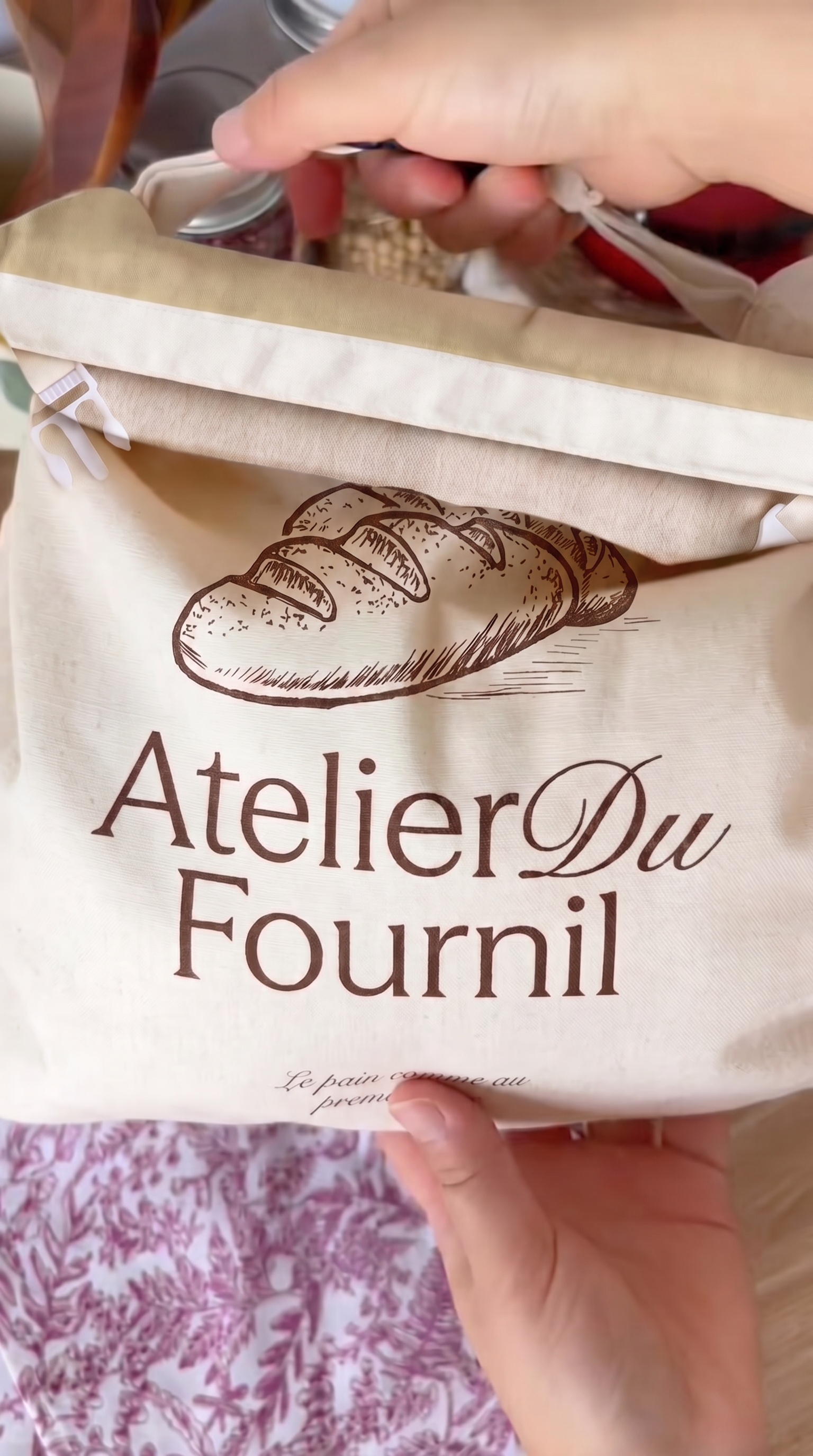 A person holding a beige cloth bread bag with 'Atelier Du Fournil' printed on it.