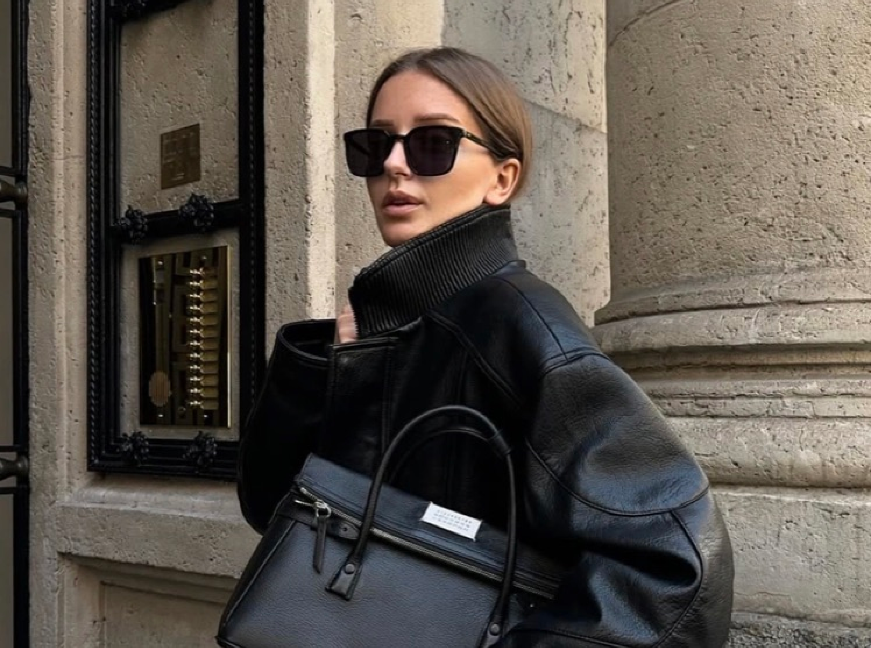 Person in black coat and sunglasses holding a handbag, standing by a stone wall.