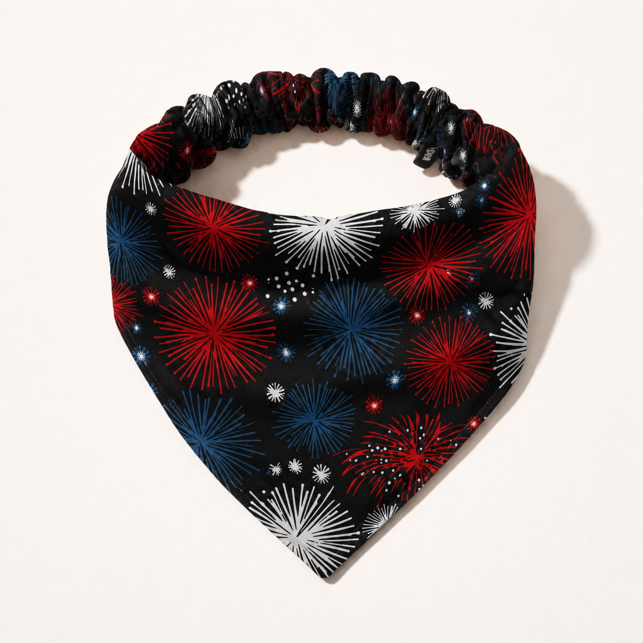 A black triangular bandana with a red, white, and blue firework print and an elastic band.