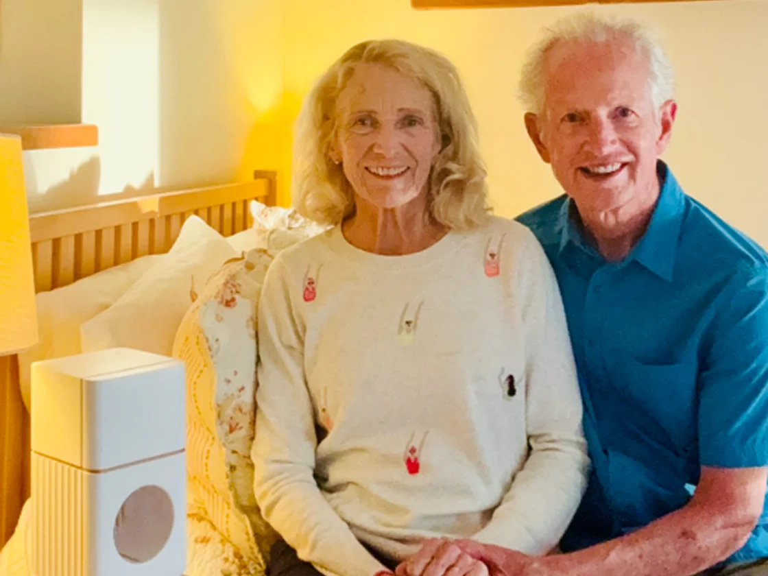 Elderly couple sitting on a bed with a lamp and device nearby.