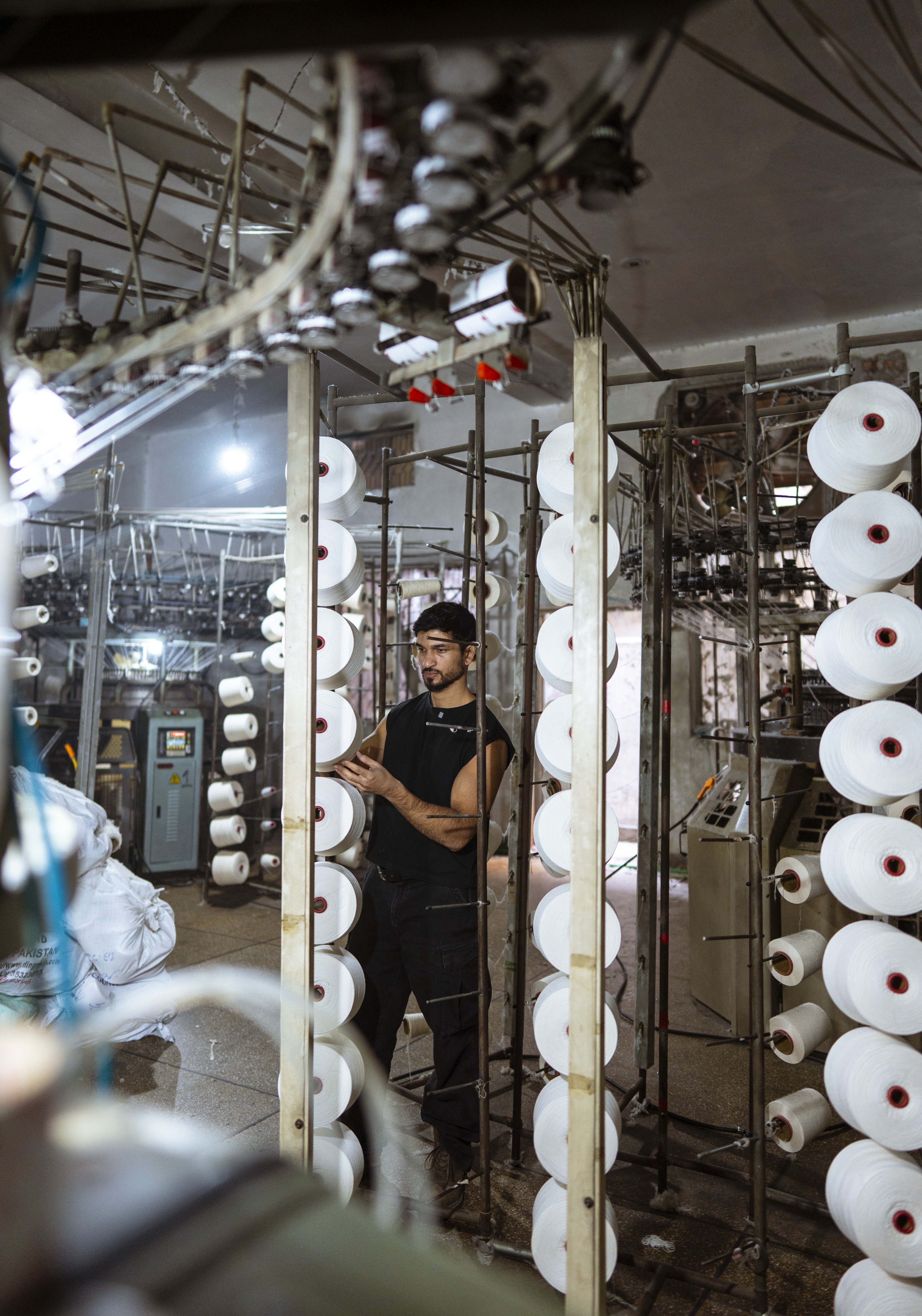 A man in a textile factory stands among industrial machines and large spools of white thread.