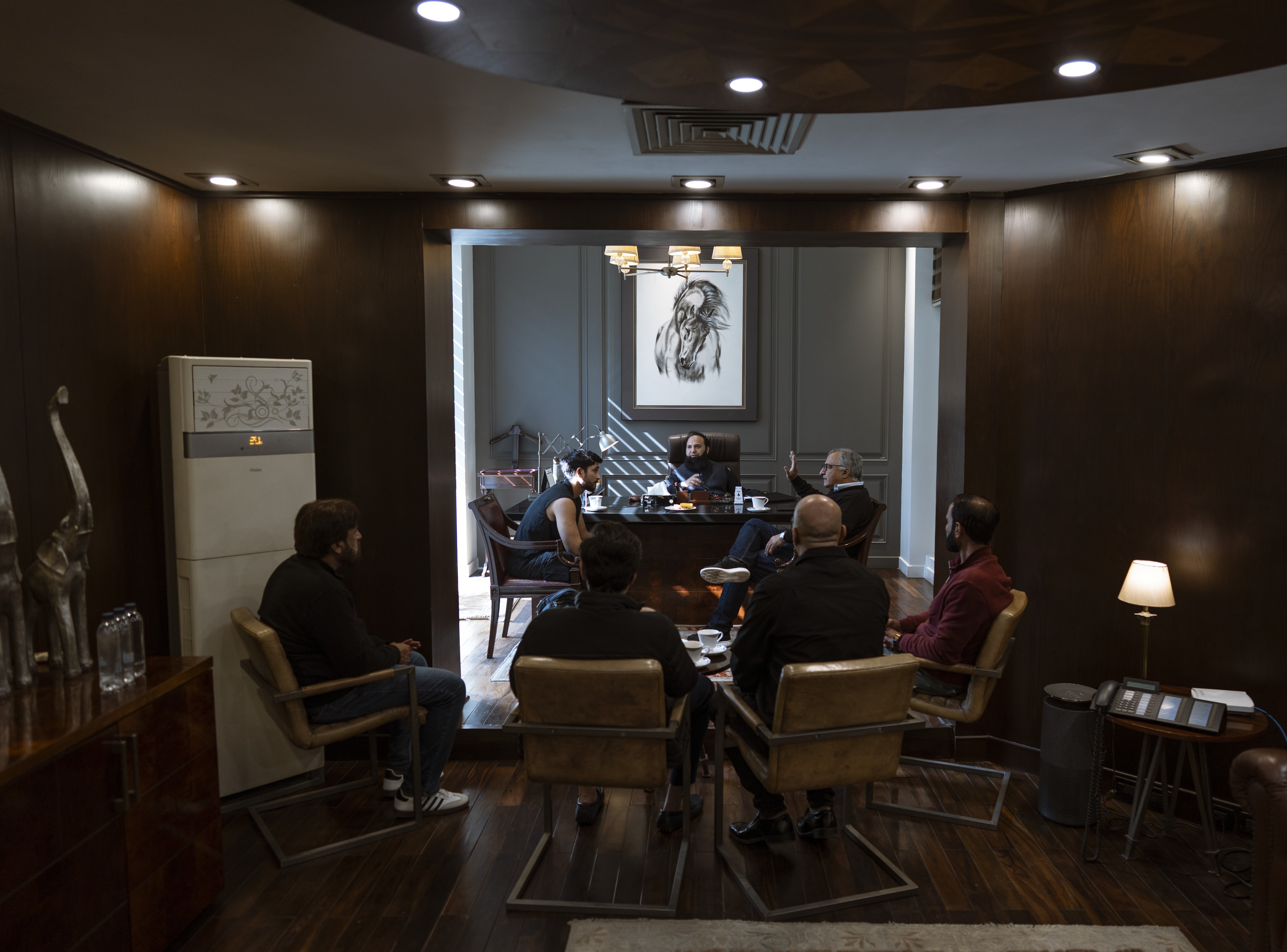 Seven men sit in chairs in a wood-paneled room, appearing to be in a business meeting.