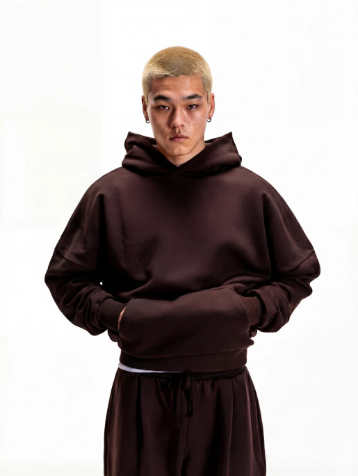 A person with short bleached hair wears a dark brown hoodie and matching pants against a white background.
