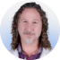A person with curly hair and a beard, wearing a white coat, against a light background.