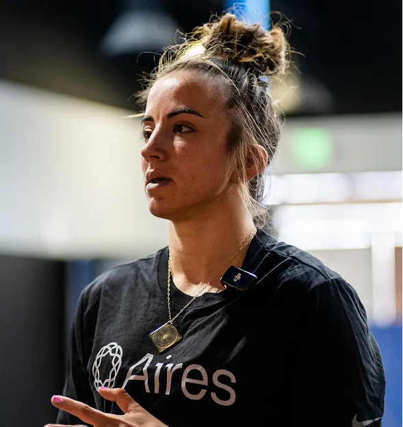 A woman with her hair in a bun, wearing a black t-shirt and a microphone, looks off-camera.