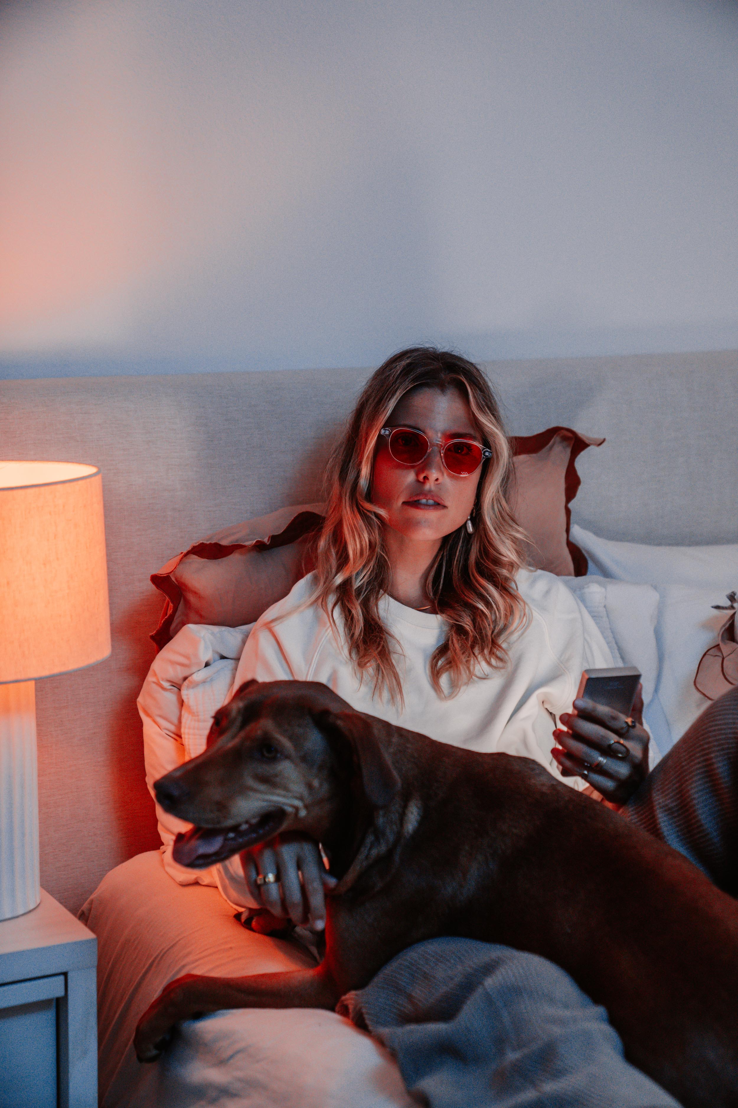 Person with sunglasses holding a phone, sitting with a dog on a bed, next to a lamp.