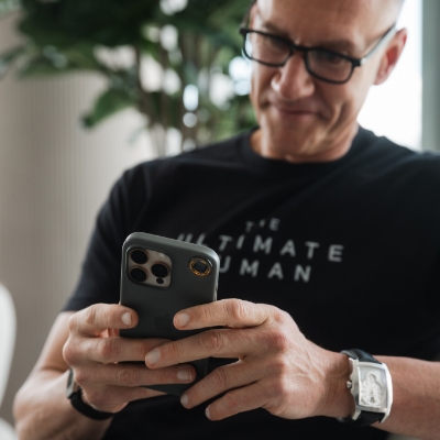 A man with glasses wearing a black t-shirt looks down at his smartphone while smiling.
