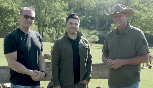 Three men stand together outdoors in a rural setting in front of a wooden fence.