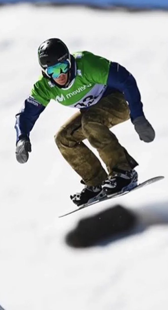 Snowboarder in mid-air wearing green and blue gear on a snowy slope.