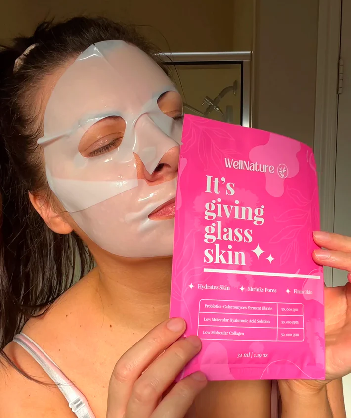 Person wearing a sheet mask holding a pink skincare product package.