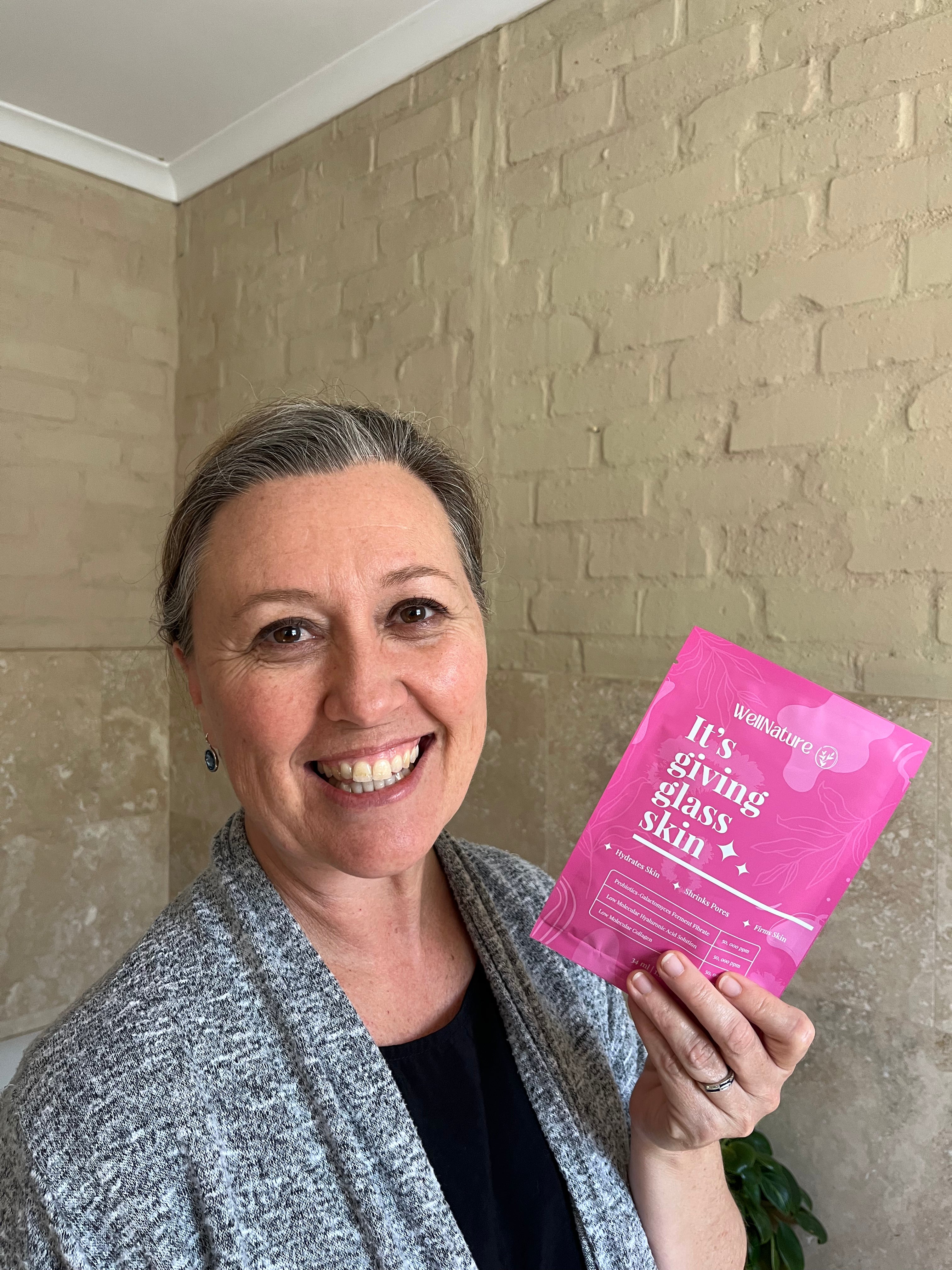 Smiling person holding a pink skincare product package against a brick wall.
