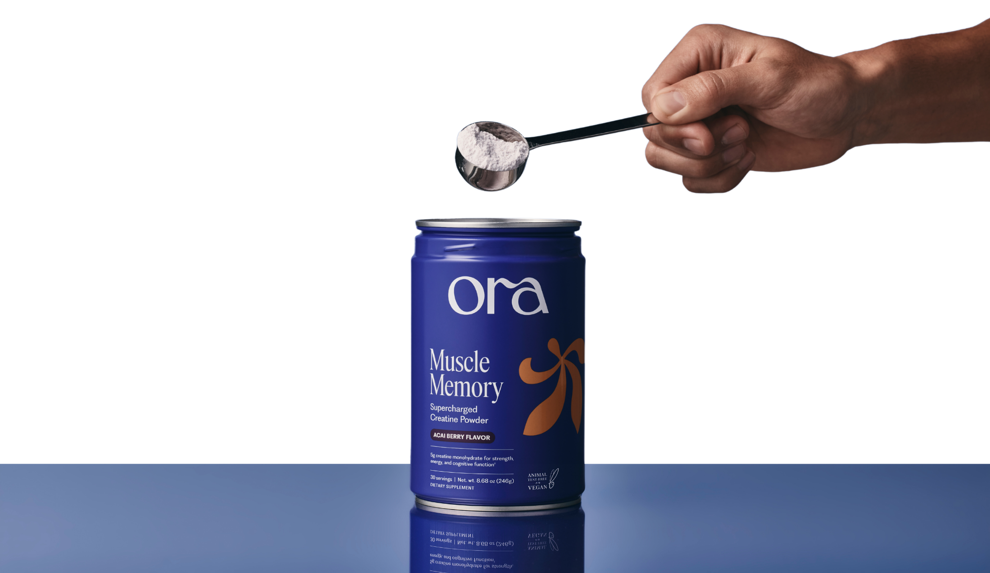 A hand holding a scoop of white powder over a blue can of Ora Muscle Memory creatine.
