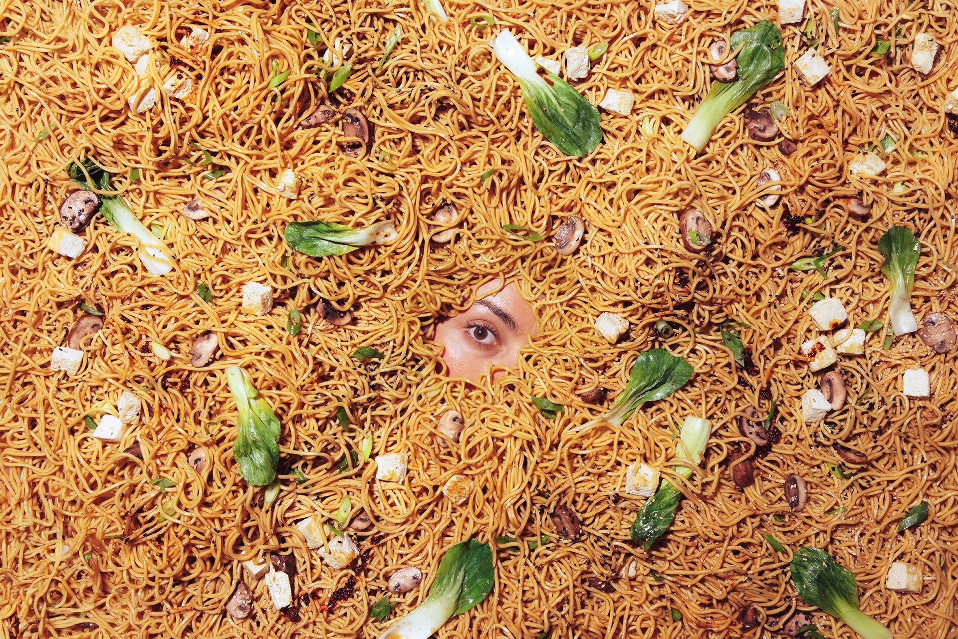A person's eye peeking out from underneath a large bed of noodles with tofu and vegetables.