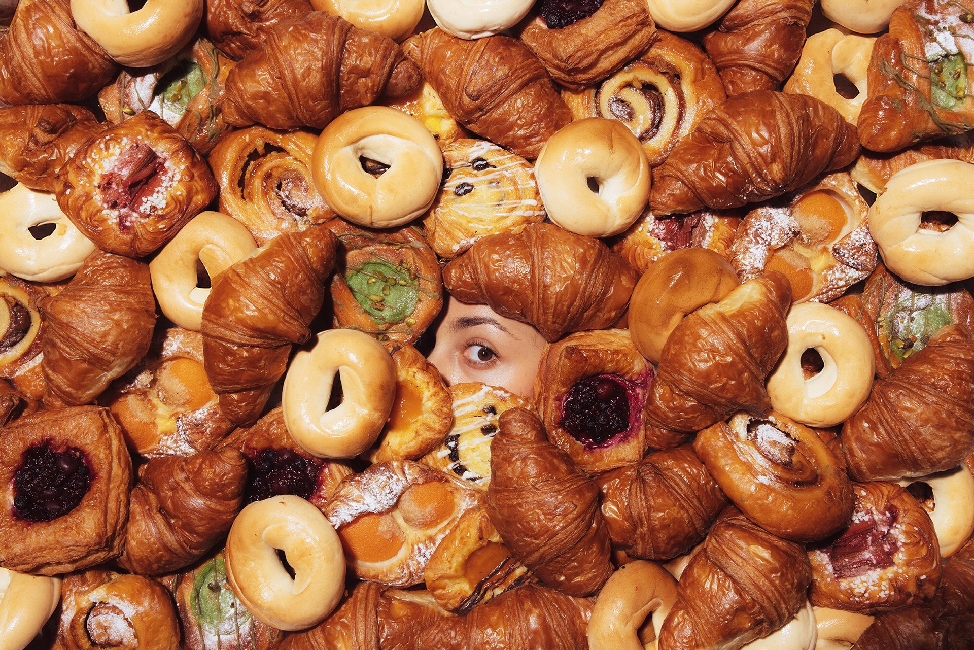 A person's eyes peek through an assortment of pastries including croissants and bagels.
