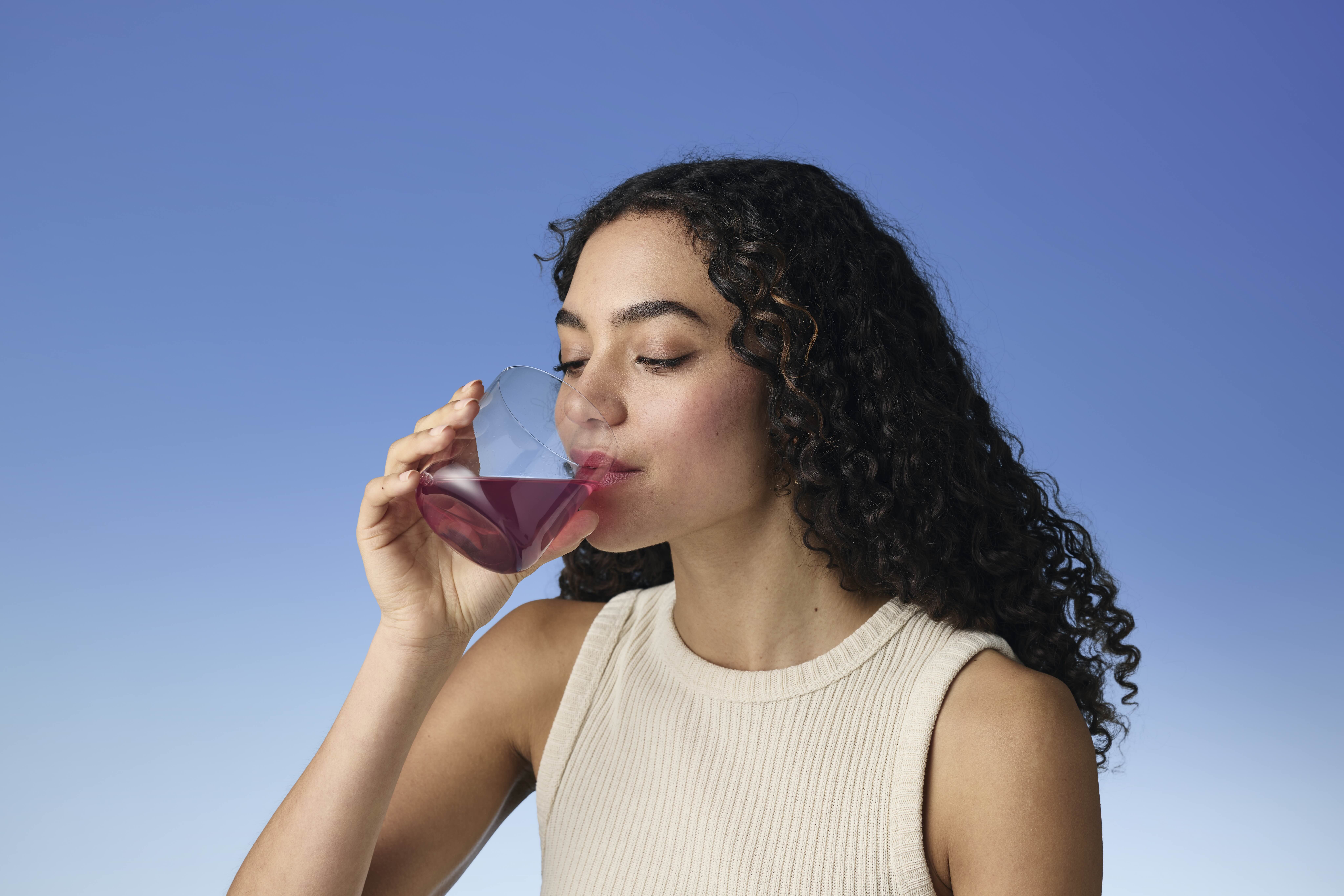 Person drinking a dark red beverage against a clear blue sky background.