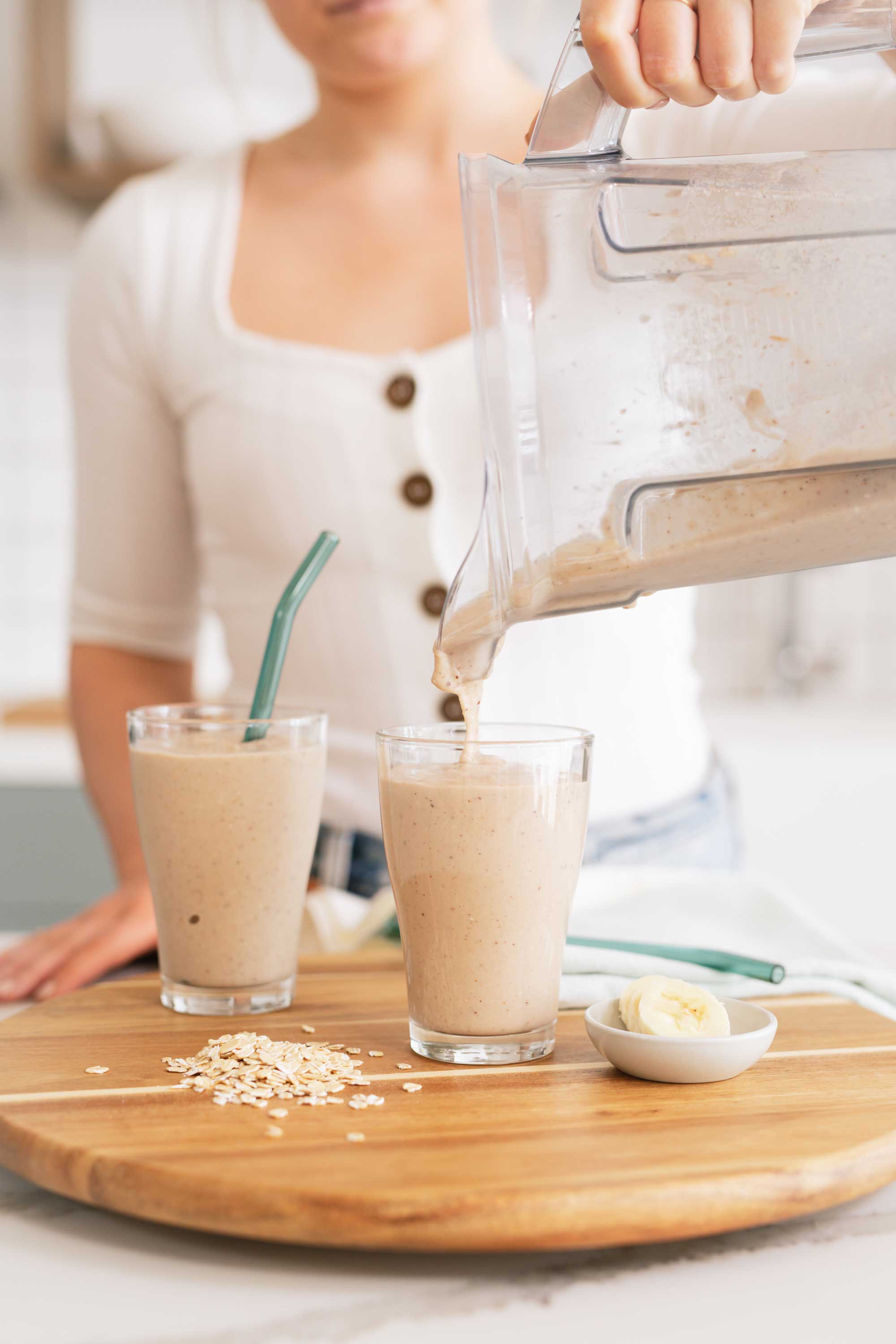 Person pouring smoothie into glass on wooden board with oats and banana slices.
