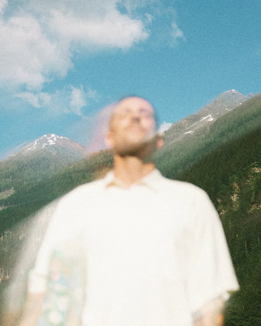 A blurry, out-of-focus person in a white shirt stands in front of a mountain range.