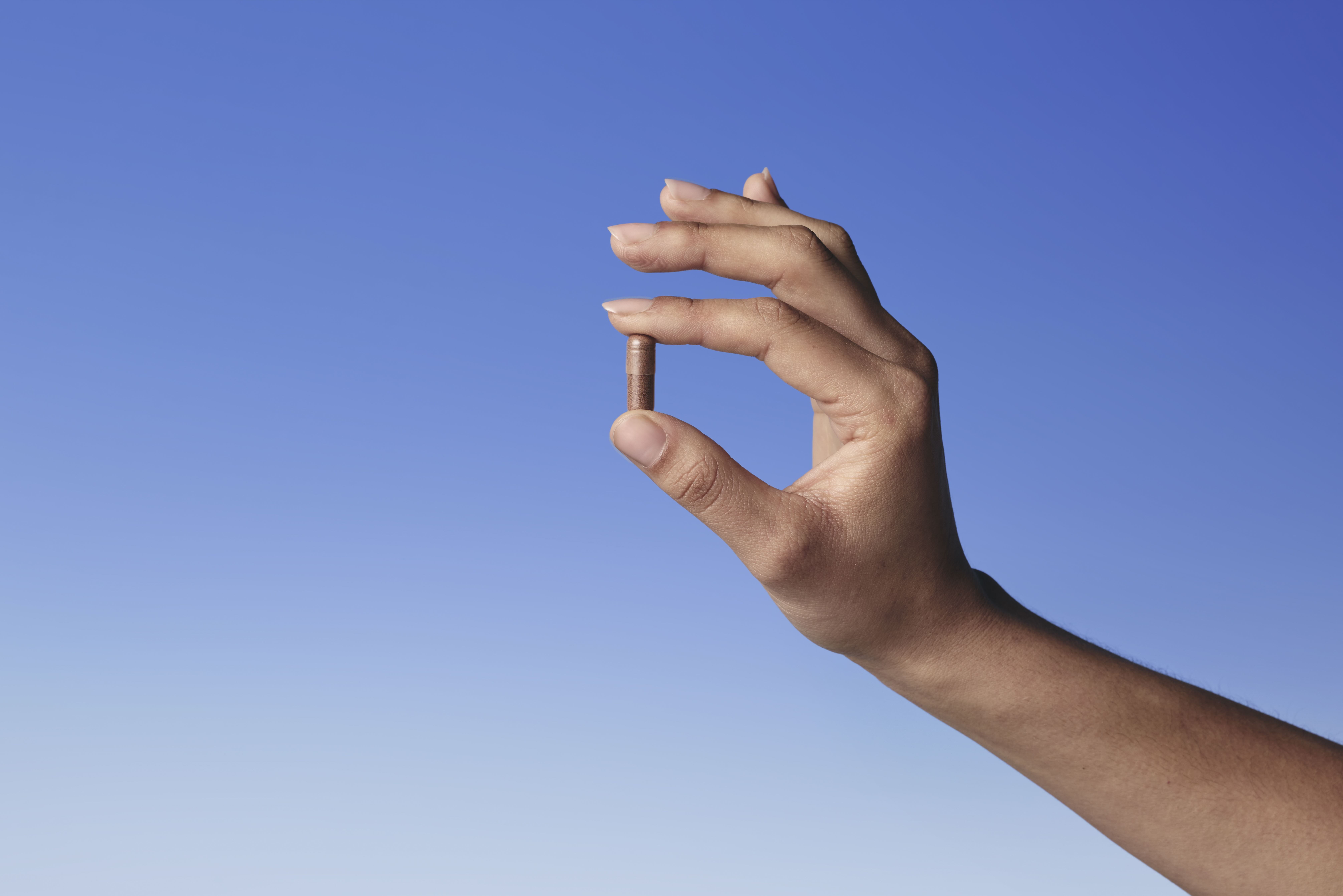 A hand holds a single brown capsule between its thumb and forefinger against a clear blue sky.