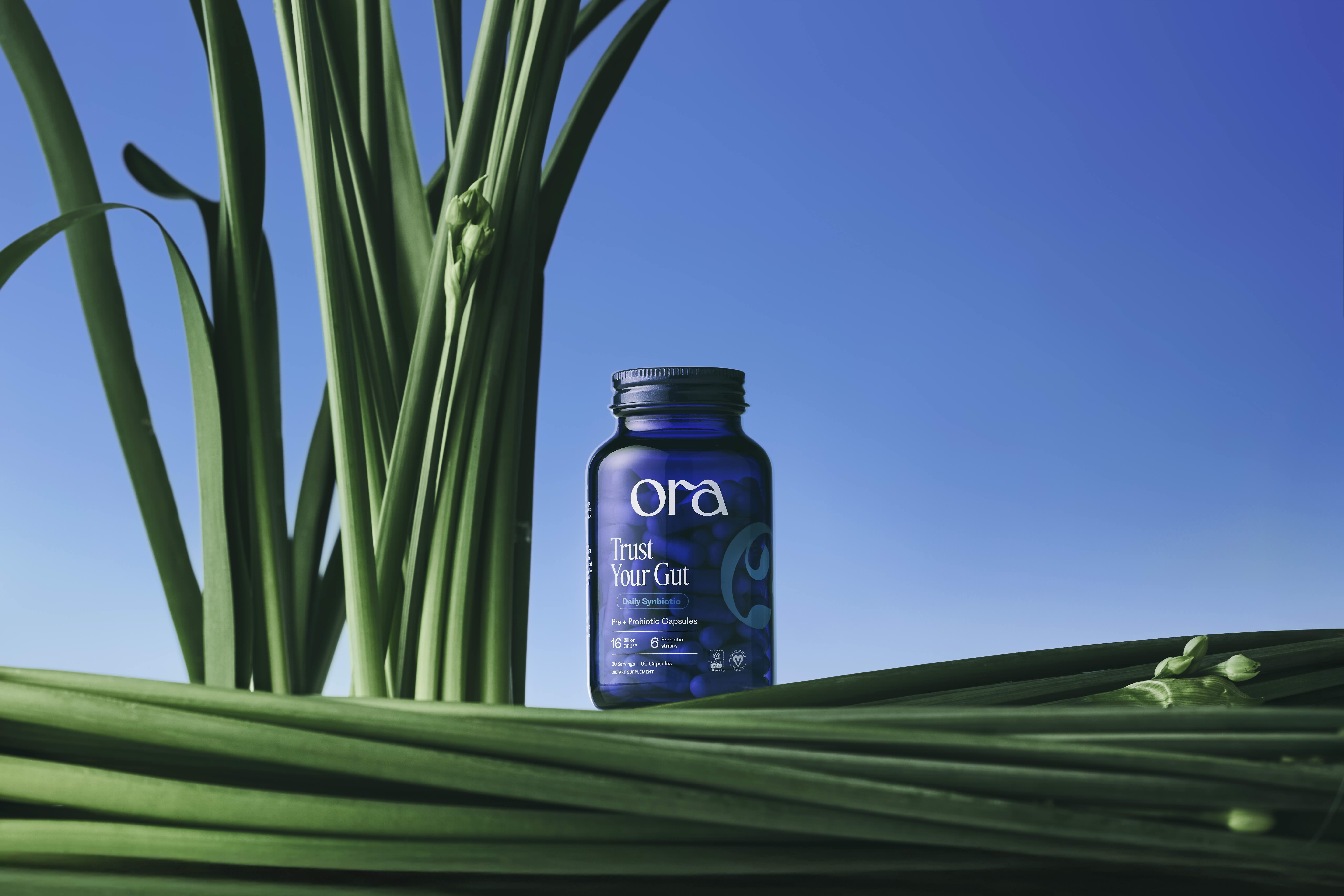 A blue bottle of Ora 'Trust Your Gut' supplements sitting amongst blades of tall green grass.